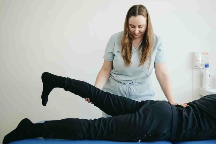 A physiotherapist assists a patient lying on an examination table in raising their leg during a rehabilitation session.