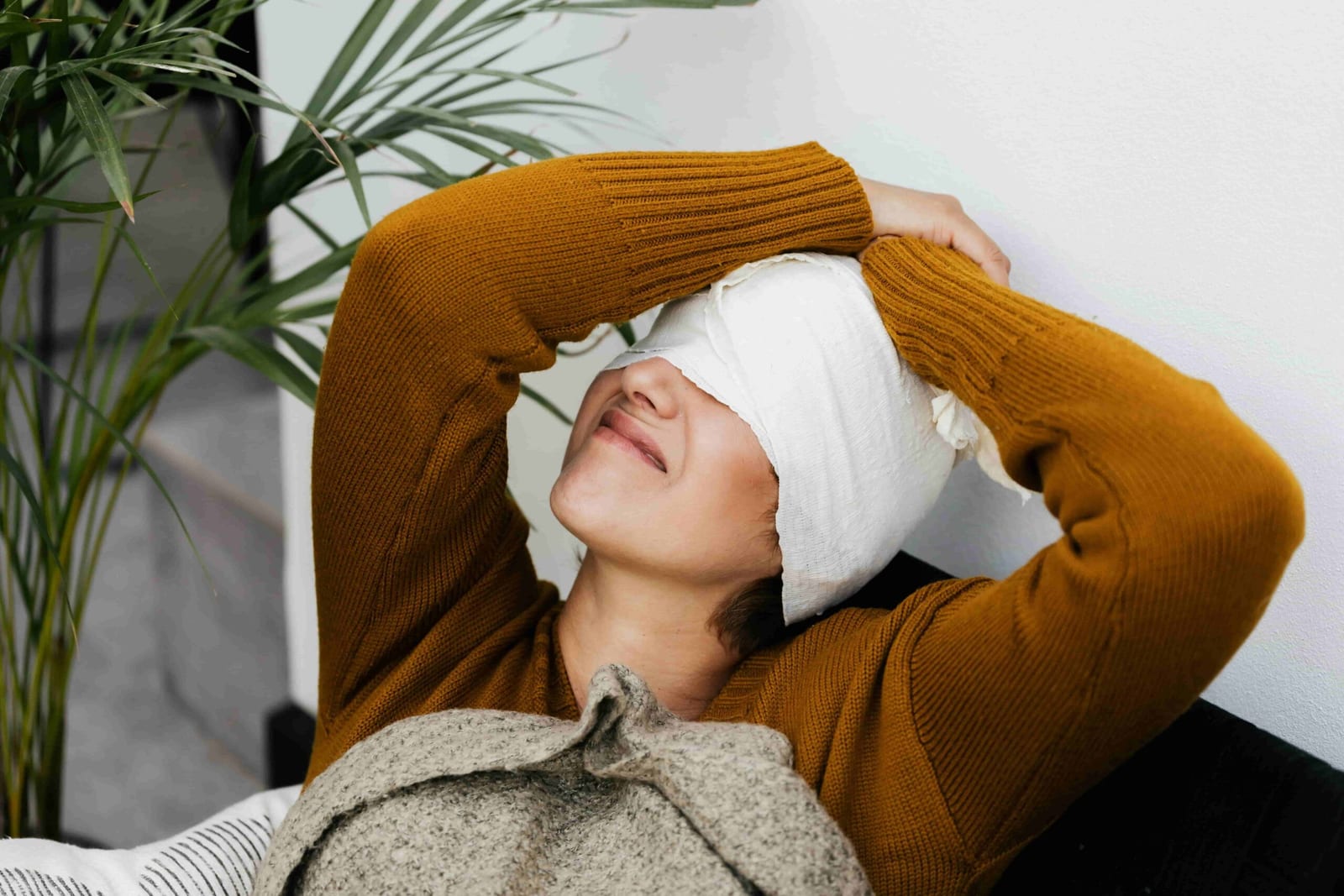 Person lying down with a bandaged head in a mustard sweater, arms over their head, resting against a pillow next to a green plant, depicting one of the physiotherapy recovery steps for concussions in North Vancouver, Lonsdale.
