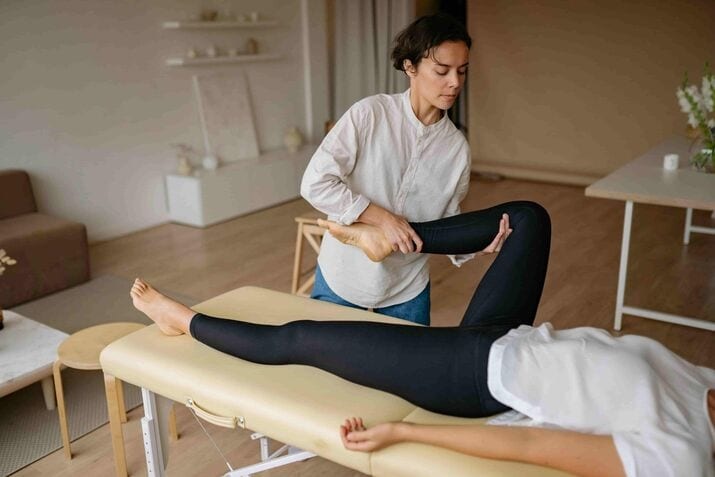 A Mobile Physiotherapy specialist assists a person lying on a treatment table by stretching their leg in a well lit room.