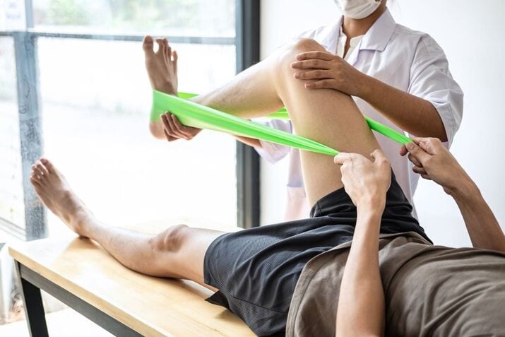 A person lying on a table uses a green resistance band for leg exercises, assisted by a masked healthcare professional who monitors their progress and rates their technique.
