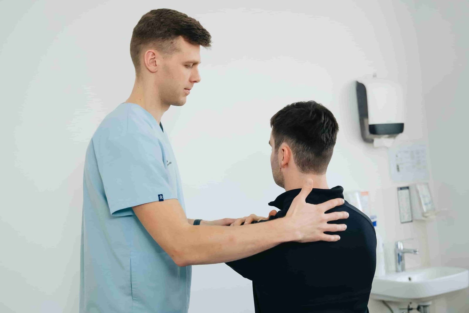A healthcare professional in scrubs examines a seated patient's shoulders in a medical office, recommending top shoulder pain physio exercises Coquitlam patients trust.
