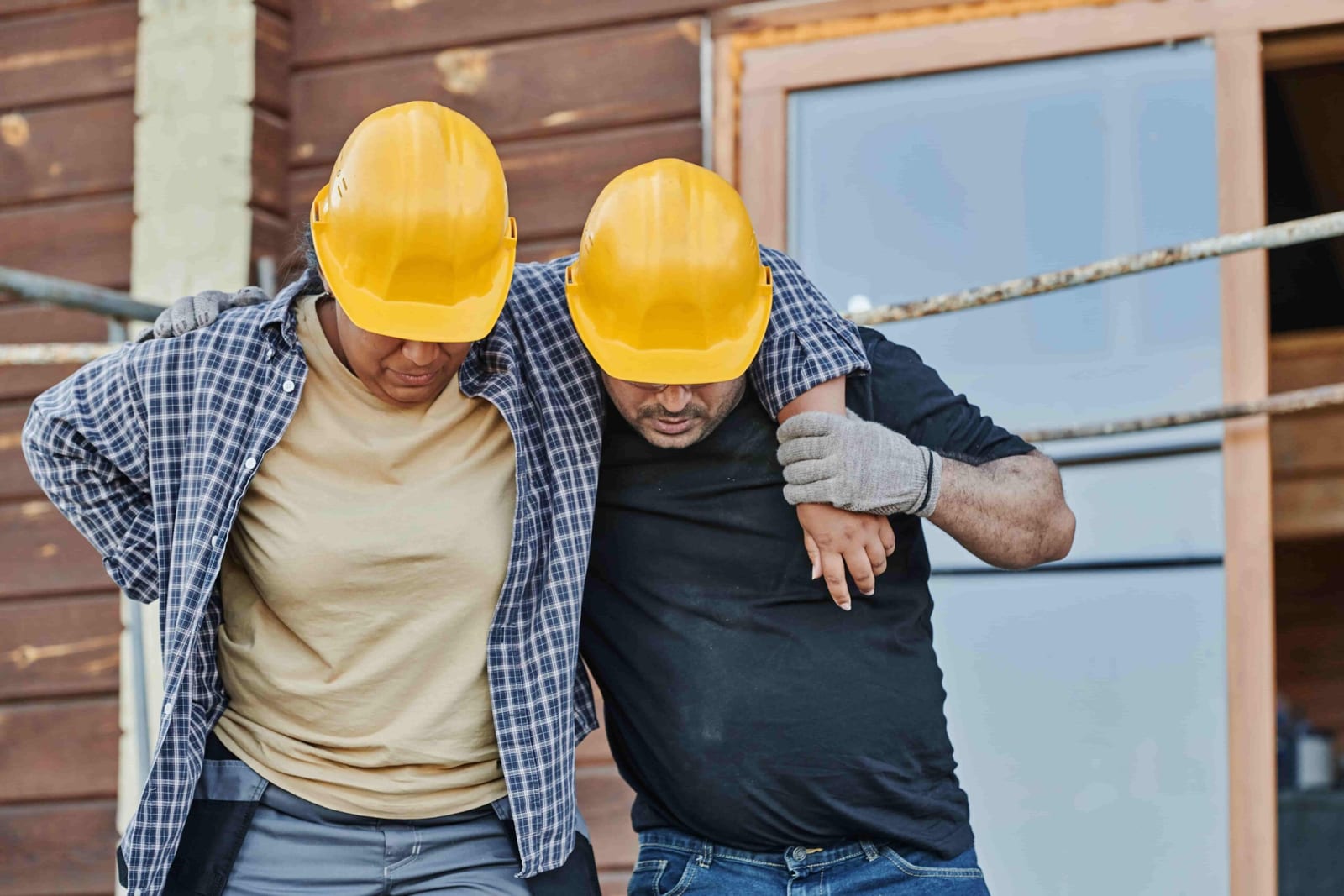 Two construction workers wearing yellow hard hats; one supports the other who appears injured, highlighting the importance of workplace injury rehab physiotherapy in North Vancouver, in front of a building with wooden walls and scaffolding.