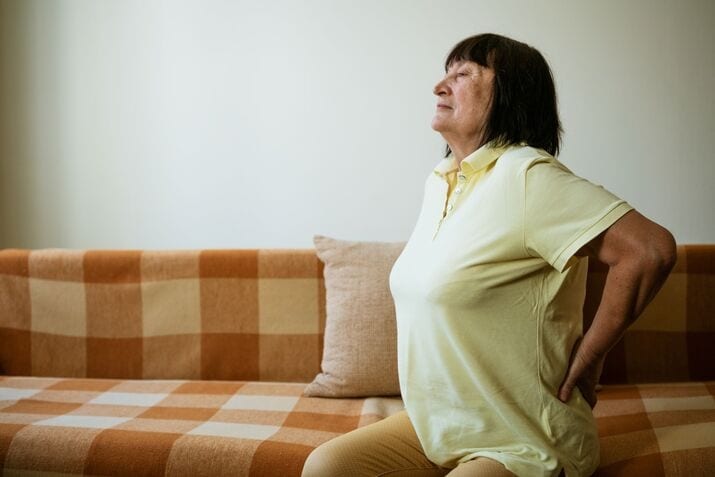 Older woman in a yellow shirt sits on a checkered sofa, holding her lower back and appearing to experience sciatica related back pain.