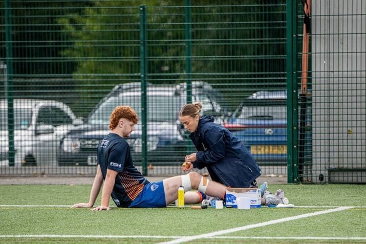 A sports player sits on artificial turf as a medic wraps his knee, providing physiotherapy for sports injury; medical supplies are spread out next to them near a fenced area.