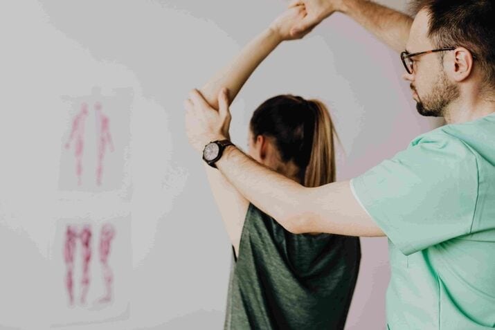 A physical therapist assists a woman in stretching her arm during a physiotherapy appointment, with anatomical diagrams visible on the wall in the background.