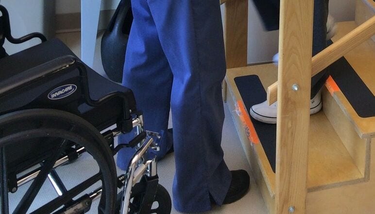 An Occupational Therapist in blue pants assists another person wearing white shoes as they walk up wooden therapy stairs next to a wheelchair.
