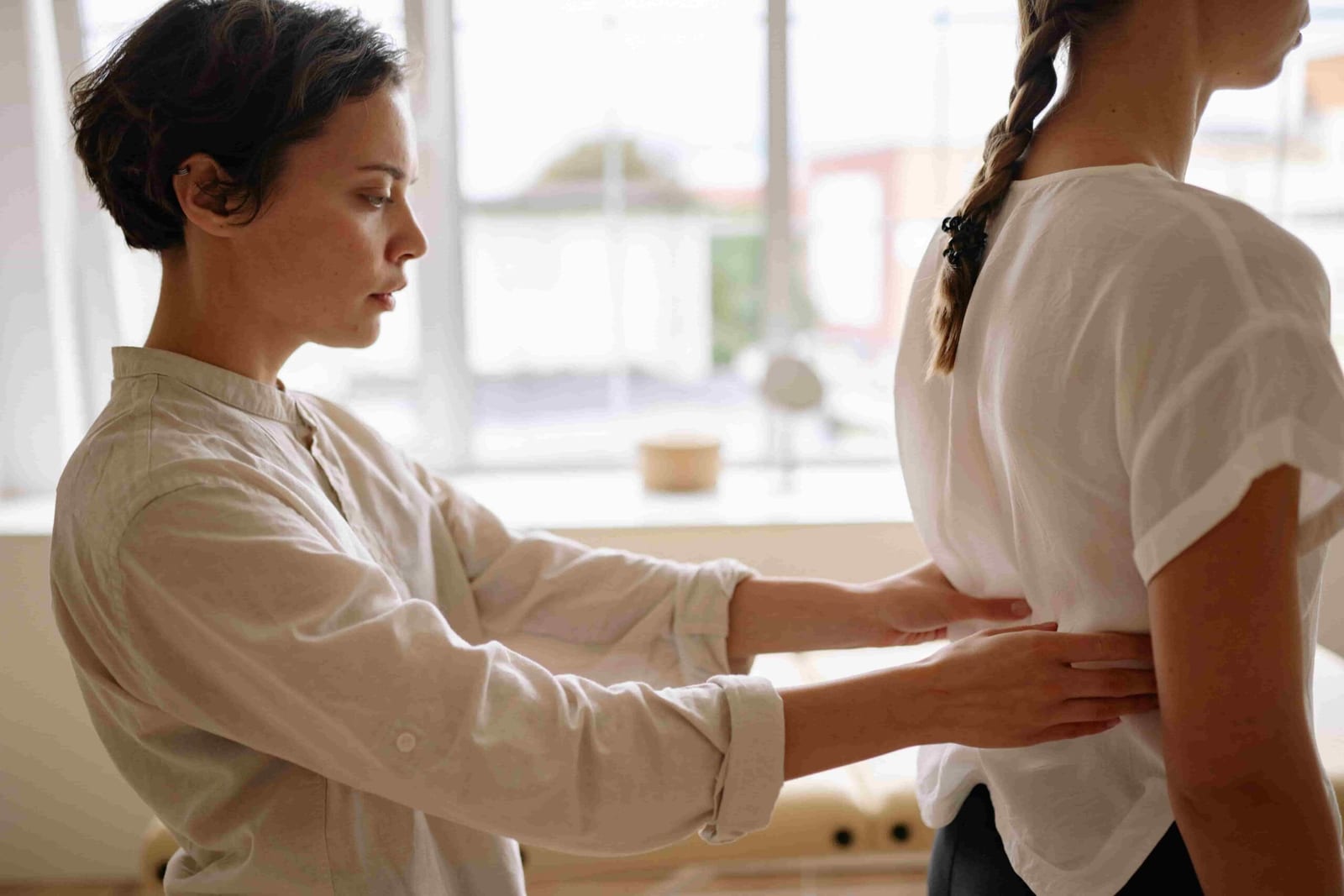 A person in a white shirt examines or adjusts another person's lower back in a bright indoor setting, reflecting the common question: Should I See a Physiotherapist or Chiropractor for My Lower Back Pain?.