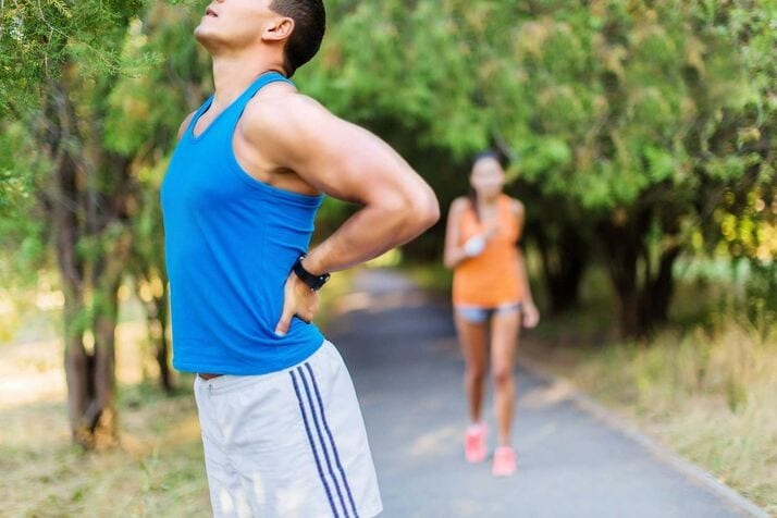Man in a blue tank top holding his lower back in discomfort, possibly due to lower back spasms, on a path with a woman in sportswear walking in the background.