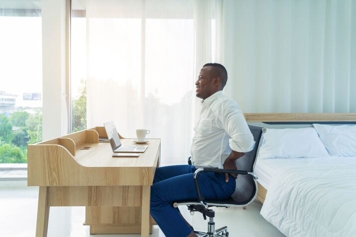 Man in business attire sitting at a desk in a bright bedroom, holding his lower back while looking at a laptop, suggesting discomfort or sciatica pain.