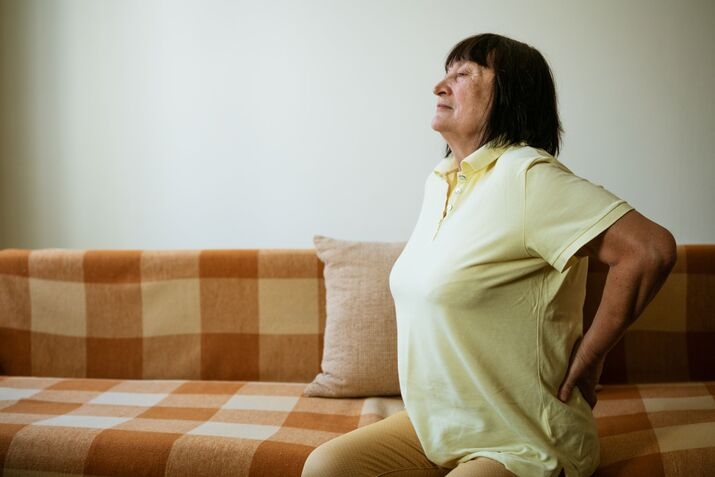 Older woman in a yellow shirt sits on a checkered sofa, holding her lower back and appearing to experience sciatica-related back pain.