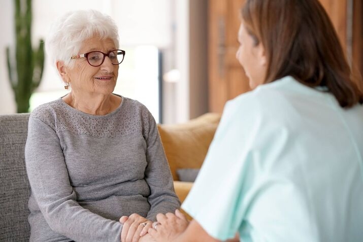 An elderly woman with white hair and glasses sits on a couch, smiling and holding hands with a woman in light blue scrubs, as they engage in stroke rehabilitation activities together.