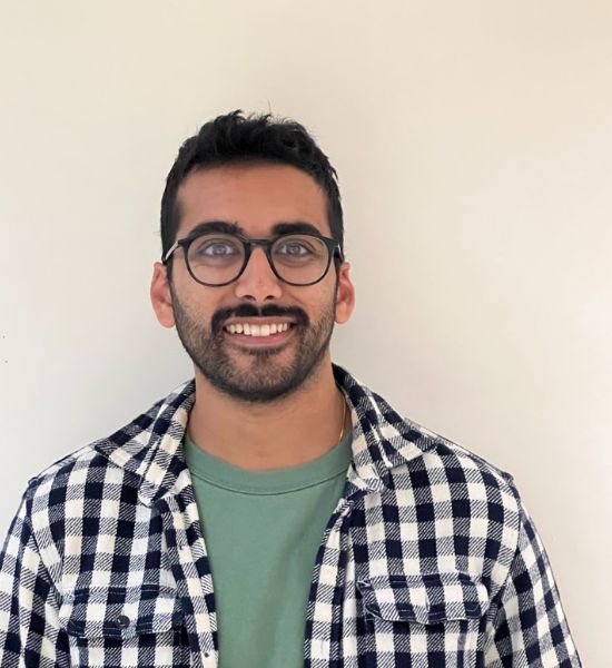 A man with glasses, a beard, and a mustache smiling at the camera, wearing a plaid shirt over a green t-shirt against a plain background. He is part of our creative team.