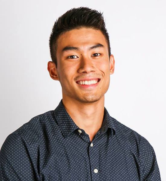 Our Team: Portrait of a smiling young man with short dark hair, wearing a dark blue patterned shirt against a plain white background.