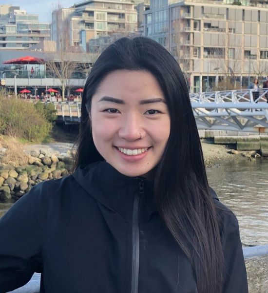 A young Asian woman smiling, wearing a black jacket, with riverfront buildings and a bridge in the background. The description provided is too brief and lacks specific content or context to accurately extract SEO keywords.