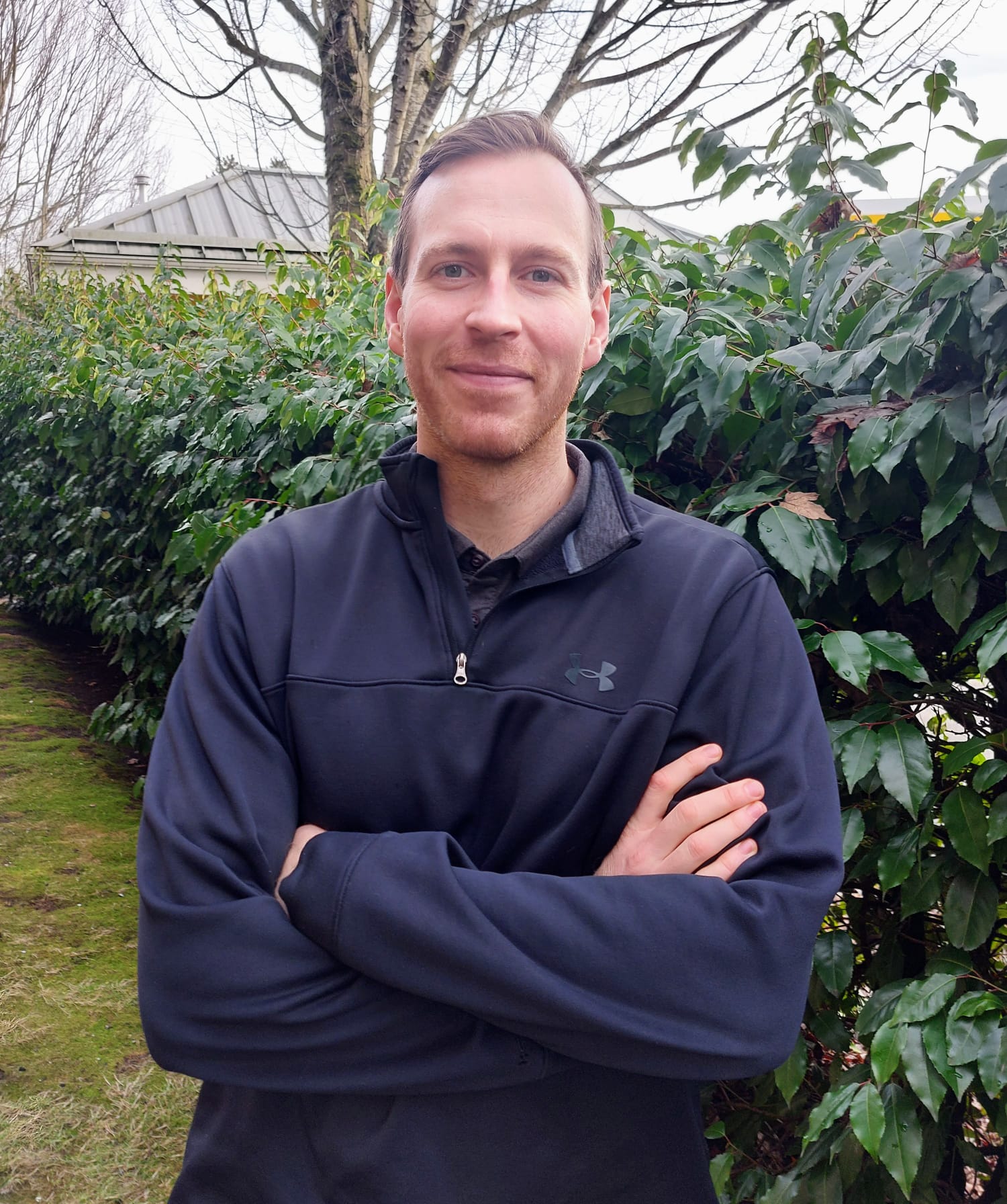 A member of our team, wearing a dark pullover, stands with his arms crossed in front of a leafy green hedge.