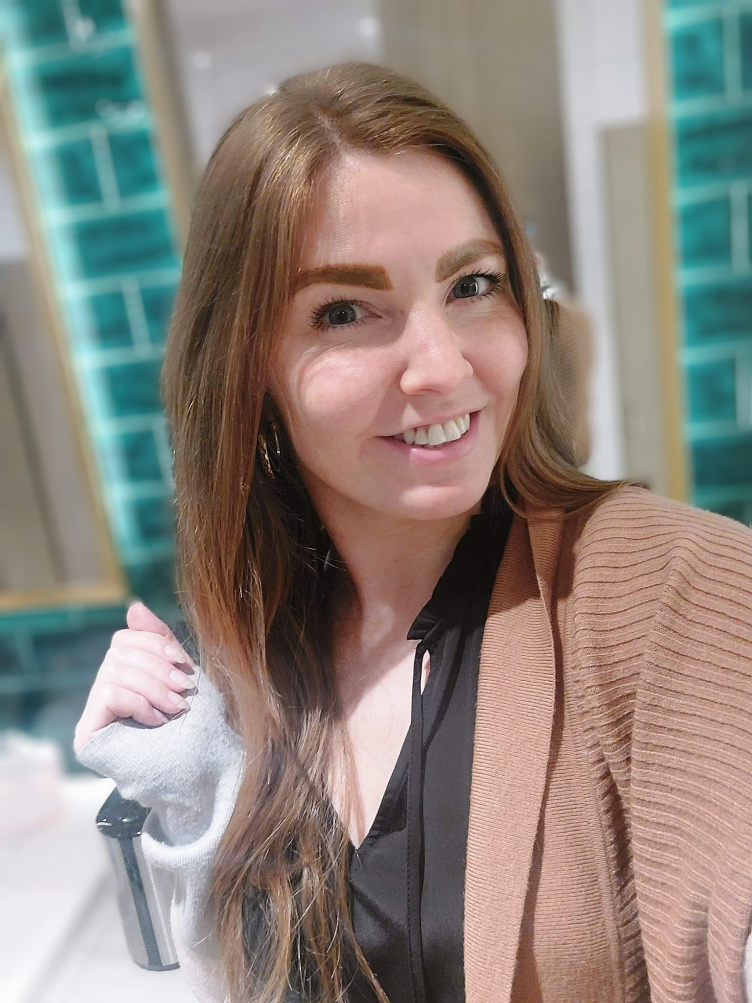 A woman with long brown hair smiles at the camera, with teal and white tiles in the background. She is wearing a brown cardigan over a black top. The setting suggests she might be part of a dedicated team, contributing her warmth and positivity to the staff members around her.