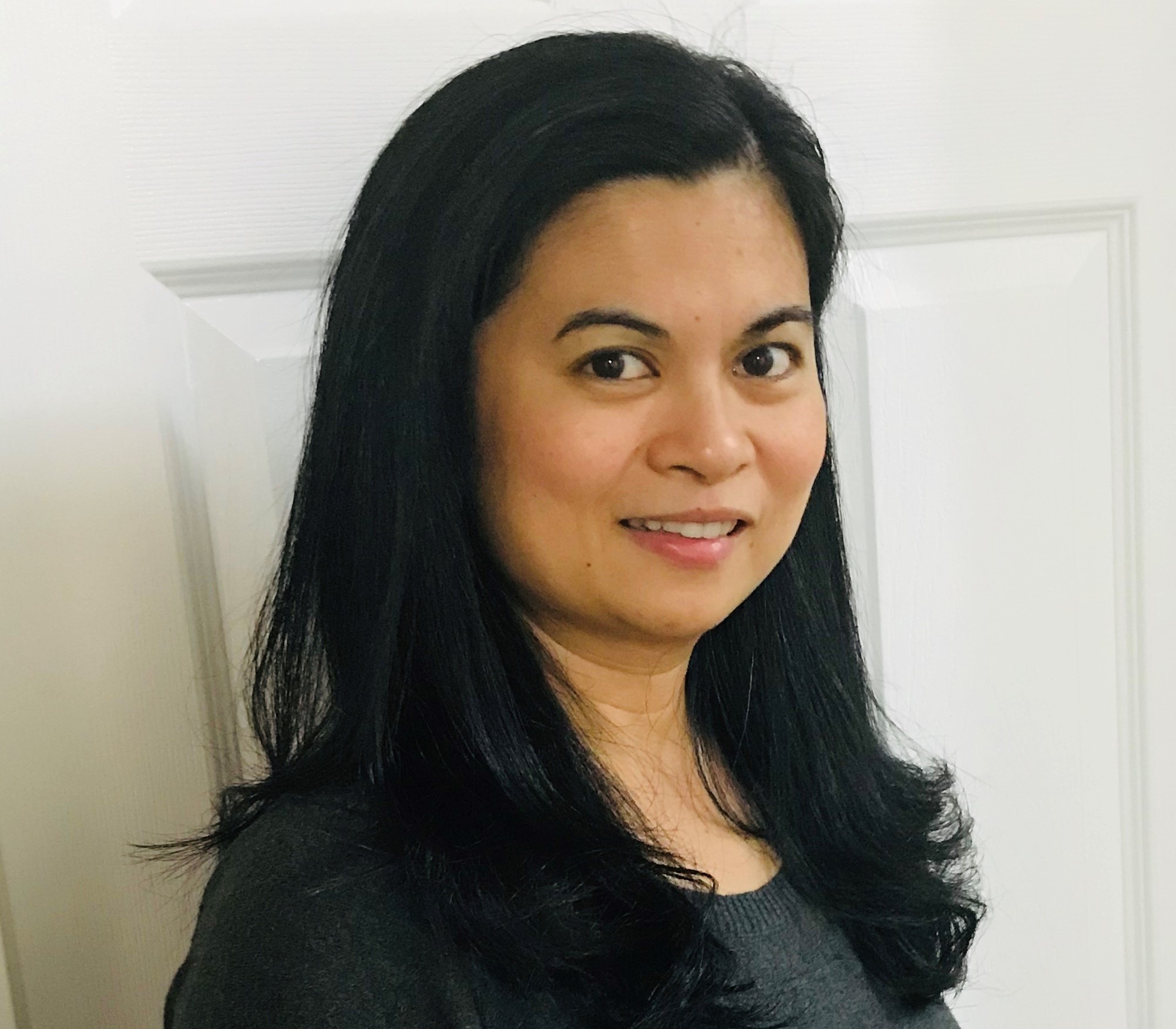 A member of our team, a woman with long black hair, wearing a dark-colored top, stands in front of a white door, looking at the camera with a slight smile.