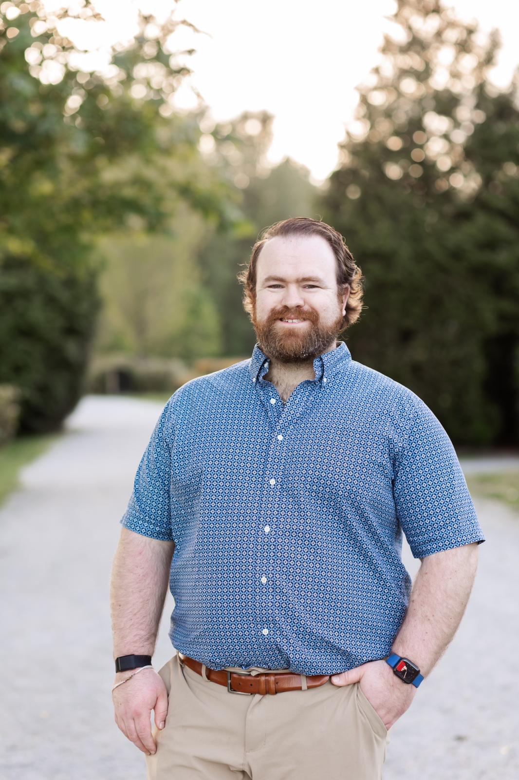 A man with a beard, wearing a blue short-sleeved shirt and beige pants, stands outdoors on a gravel path lined with trees in Coquitlam. He has his left hand in his pocket and is smiling at the camera.