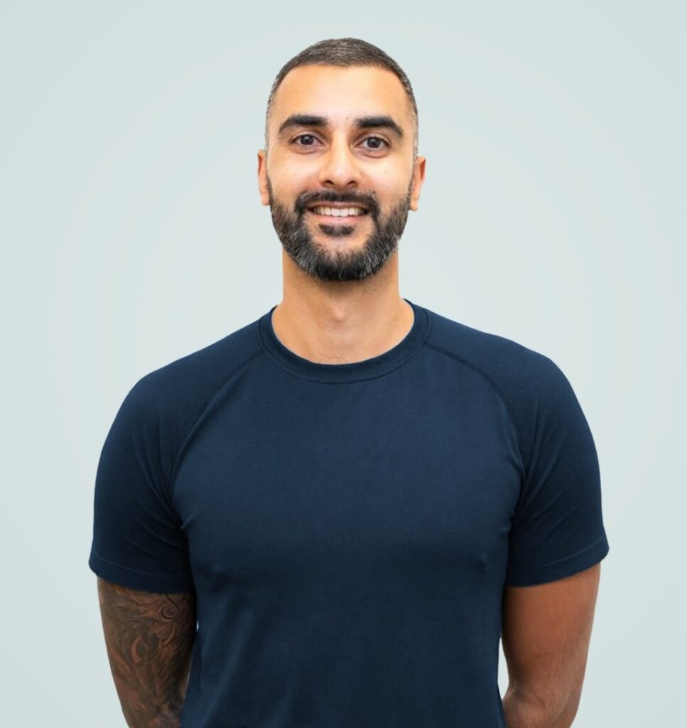 A person with short hair and a beard, wearing a navy blue t-shirt, stands smiling against a light grey background, representing our team.