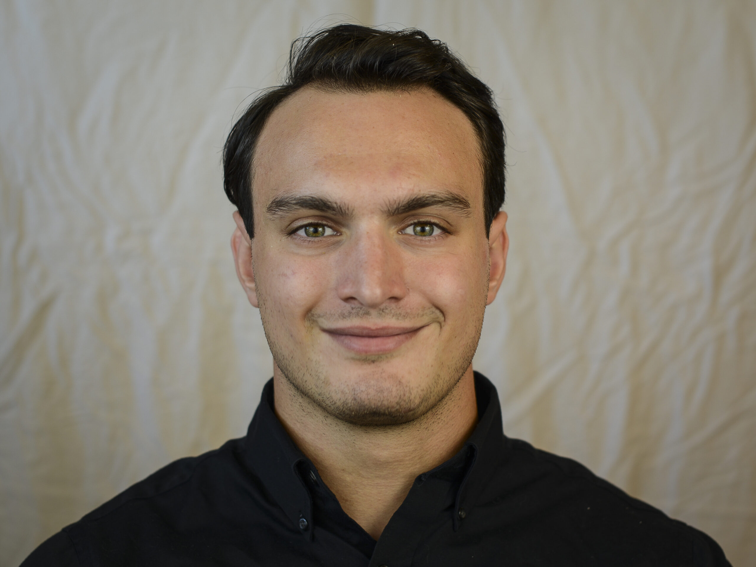 Portrait of a man with short dark hair, wearing a black collared shirt, against a light-colored fabric backdrop. Part of Our Team series.