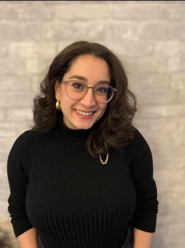 Person with shoulder-length dark hair, glasses, and a black turtleneck, smiling against a light gray brick wall background. A perfect addition to our team.