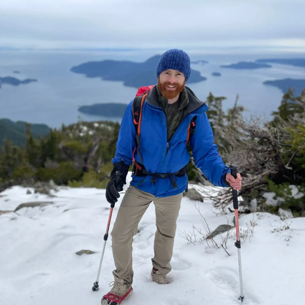A person in a blue jacket and knit hat stands on a snowy mountain slope with hiking poles, overlooking a scenic view of water and islands, reminiscent of an adventure by Thomas Scarr.