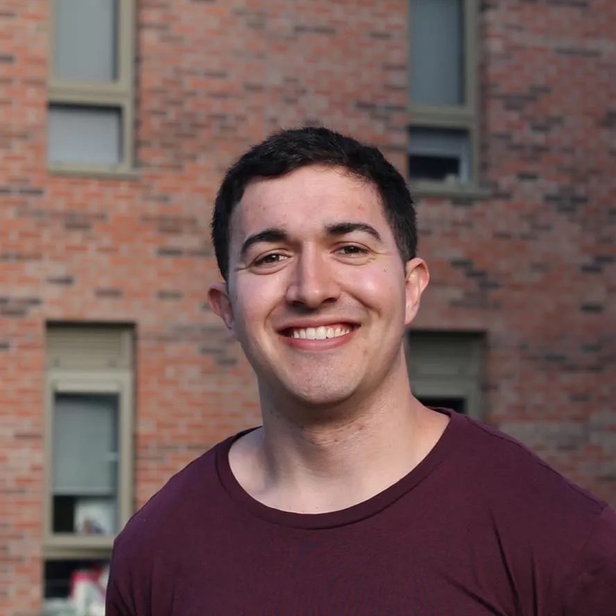 Thomas Scarr, a young man with short dark hair, wearing a maroon shirt, smiles in front of a brick building with several windows.