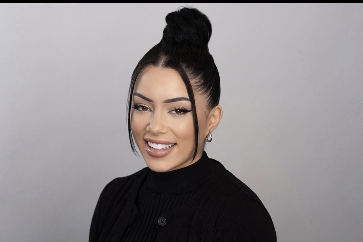 With long black hair in a bun and hoop earrings paired with a black sweater, she exudes confidence and warmth. Her smile suggests a passion for Active Rehab, radiating positivity against the gray background.