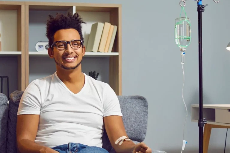 A man sits on a gray couch in a modern North Vancouver living room, receiving an IV infusion with the stand and bag beside him.