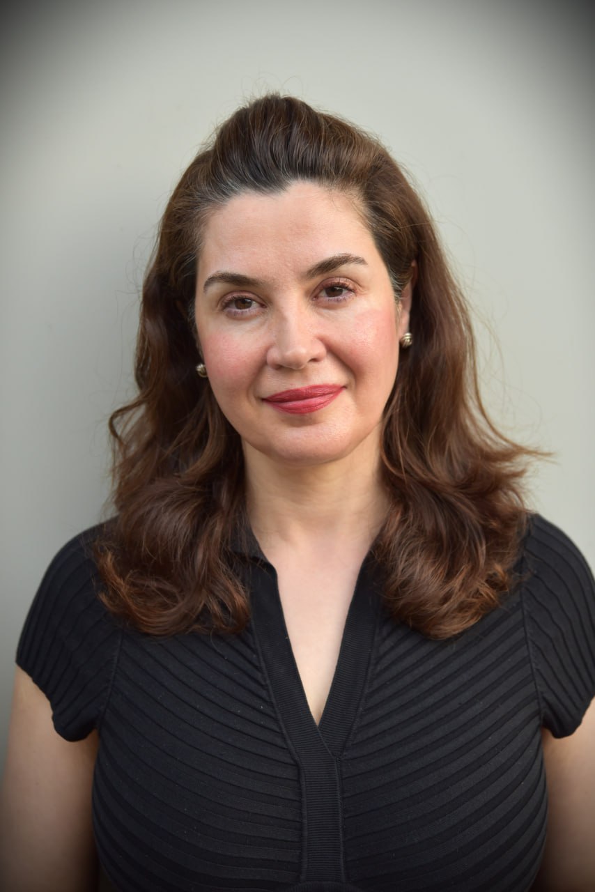 Joan Eustaquio, with wavy brown hair, wears a black short-sleeved top as she stands before a plain light-colored background, gazing at the camera with a neutral expression.