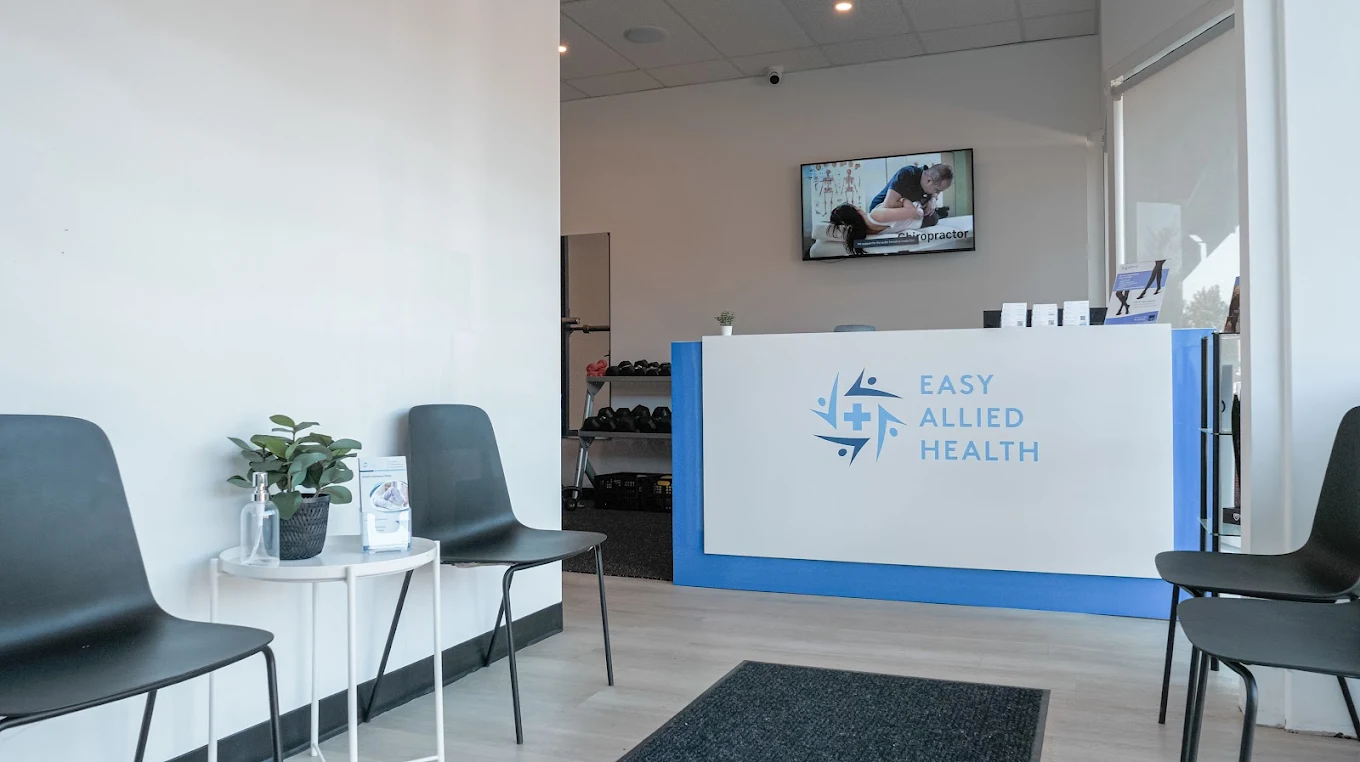 Reception area of Easy Allied Health clinic with black chairs, small white tables, a plant, and a counter with the clinic’s logo; TV on the wall in the background.