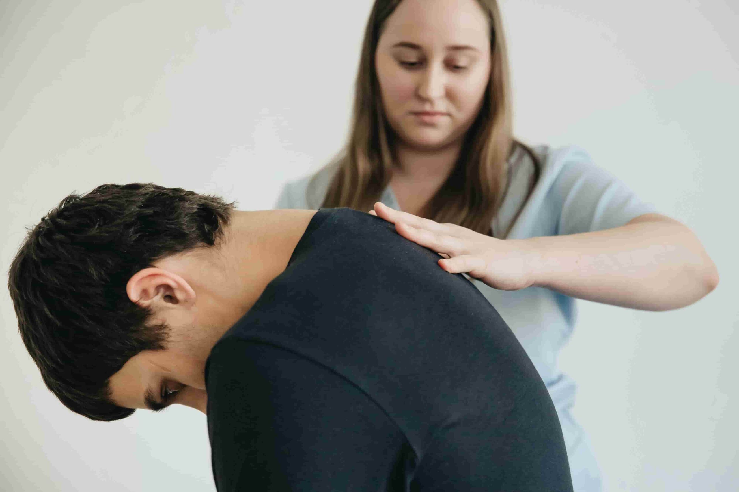 A woman in medical attire examines or treats a man's upper back and shoulder—possibly addressing pain caused by poor posture—as he sits with his head and shoulders bent forward.