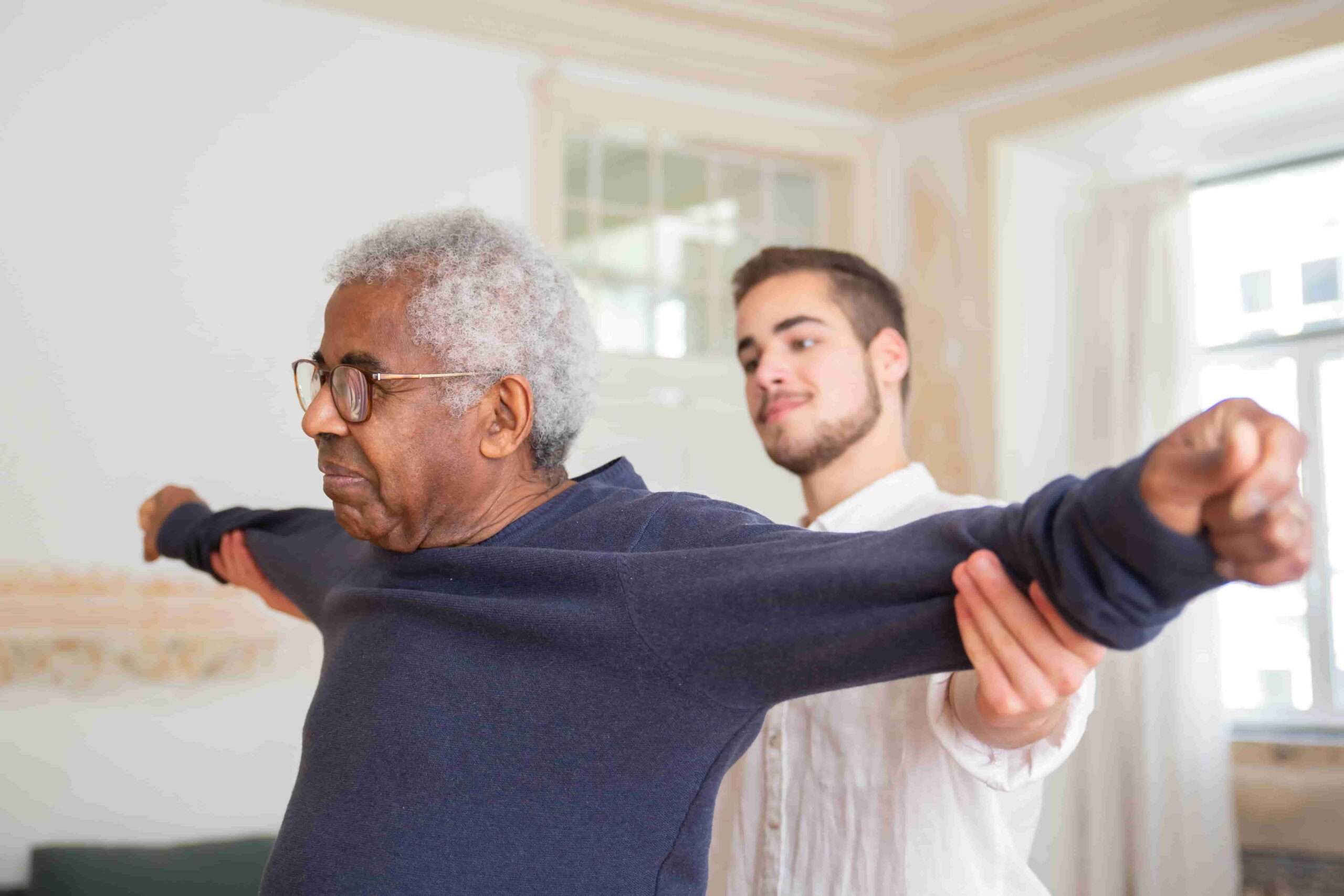 A younger man assists an older man in stretching his arms in a bright room, demonstrating physiotherapy for seniors in Coquitlam targeting arthritis pain.