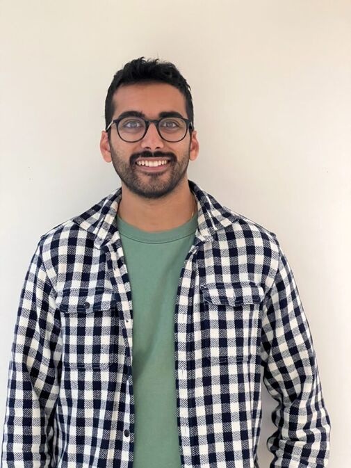 A man with short dark hair, a beard, and glasses is standing against a plain white background, wearing a green shirt and a black-and-white checkered jacket.
