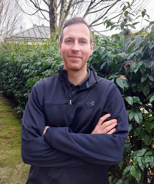A man stands outdoors with arms crossed, wearing a black zip-up jacket, in front of a leafy hedge and trees.
