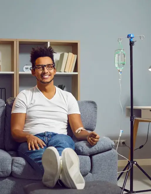 A man sits on a couch in his living room receiving IV Therapy North Vancouver, with an IV drip stand beside him.
