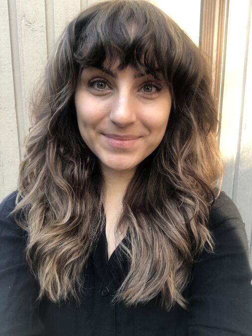 A woman with long, wavy brown hair and bangs smiles at the camera while standing in front of a light-colored wall.