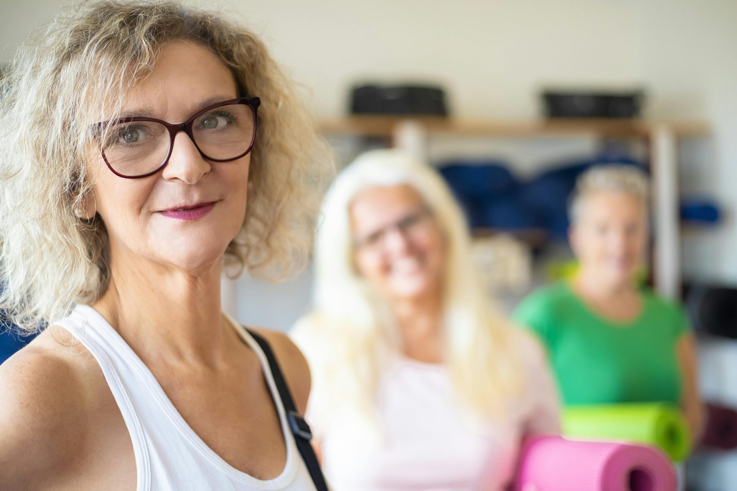 Three women, each holding yoga mats, stand indoors. The woman in front with curly hair and glasses is in focus; the two women behind her are blurred, suggesting a group physiotherapy seniors class.