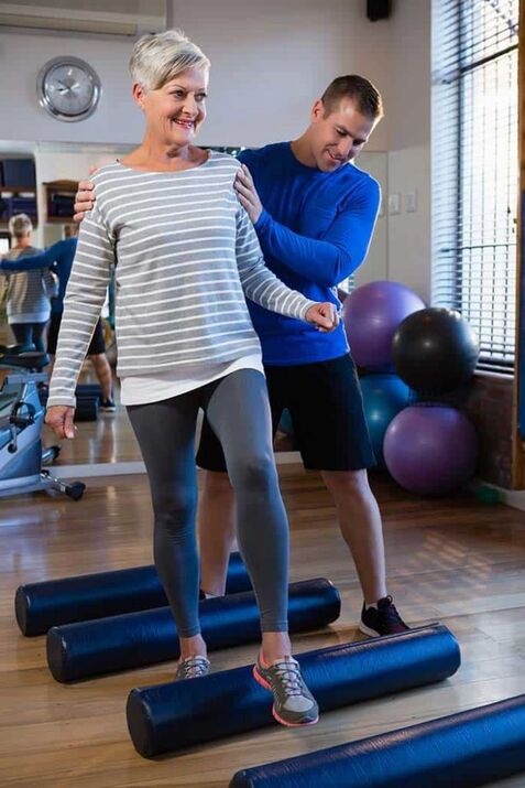 A woman practices balance by stepping on foam rollers in a gym while a trainer assists her, demonstrating techniques often used in geriatric therapy.
