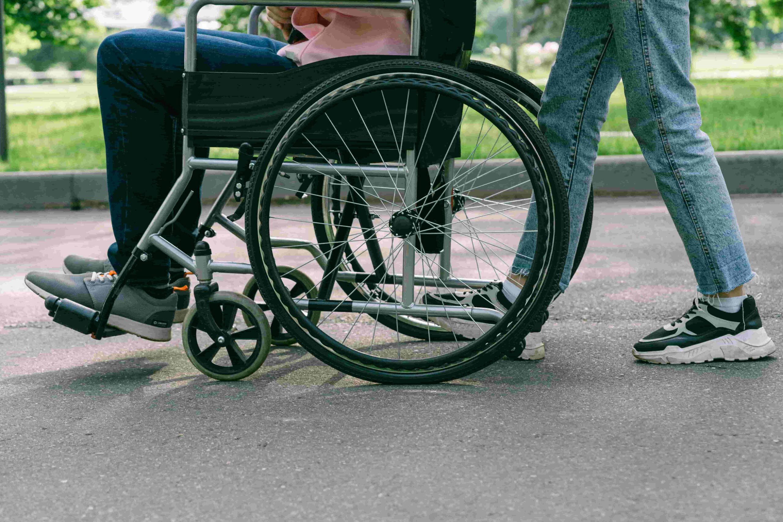 A person in a wheelchair is being pushed by another on an outdoor path, possibly as part of post-surgery physio.