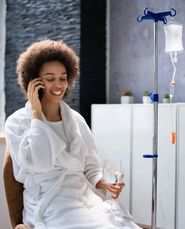 A woman in a white robe sits on a chair, talking on the phone and holding a glass of water, next to an IV Therapy Coquitlam drip stand.