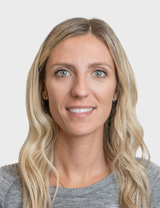 A woman with long blonde hair and light eyes, wearing a gray top, is facing the camera against a plain light background.