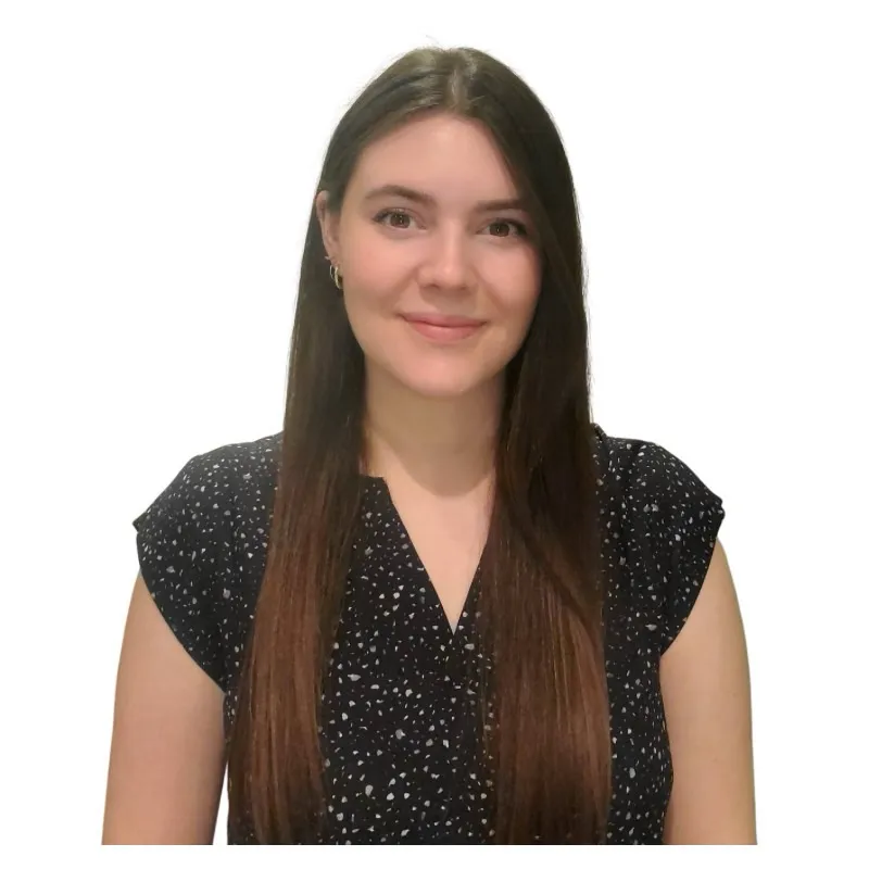 Katrina Parsley, a woman with long straight brown hair, wears a black short-sleeve blouse with white speckles, smiling and facing the camera against a plain white background.