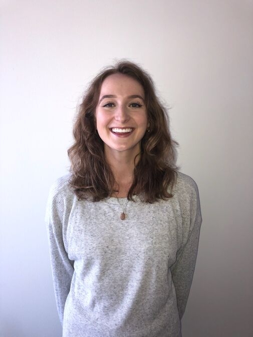 A woman with wavy brown hair, wearing a light gray sweater and a necklace, stands smiling in front of a plain white wall.
