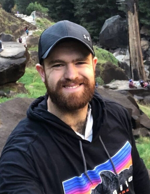 Bearded man wearing a black cap and hoodie smiles at the camera outdoors in Coquitlam, with rocks, grass, and trees in the background.