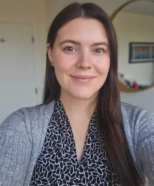 A woman with long brown hair, wearing a patterned blouse and gray cardigan, smiles at the camera indoors at a chiropractor North Vancouver office, with a mirror and shelves in the background.