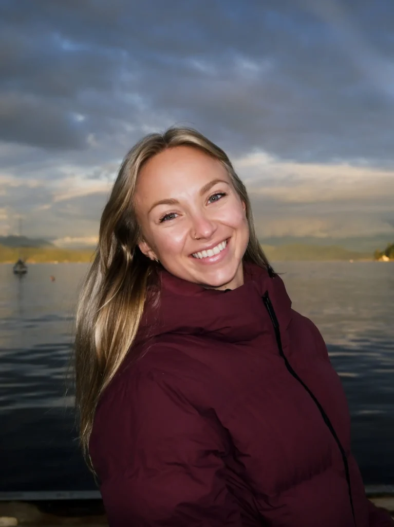 Luke Warner, with long blonde hair and a maroon jacket, smiles while standing in front of a calm lake with mountains and a cloudy sky in the background.