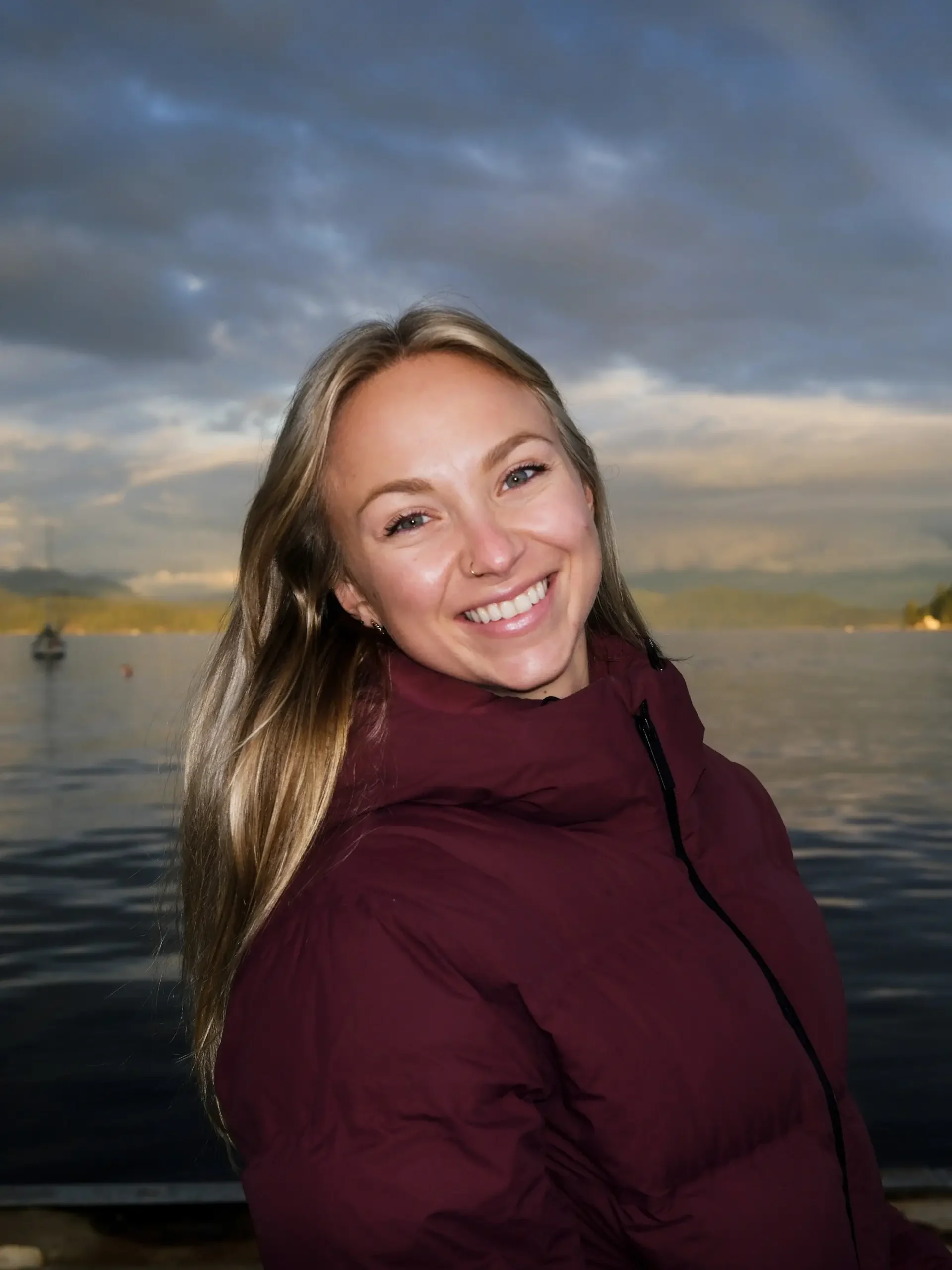 Luke Warner, with long blonde hair and a maroon jacket, smiles while standing in front of a calm lake with mountains and a cloudy sky in the background.