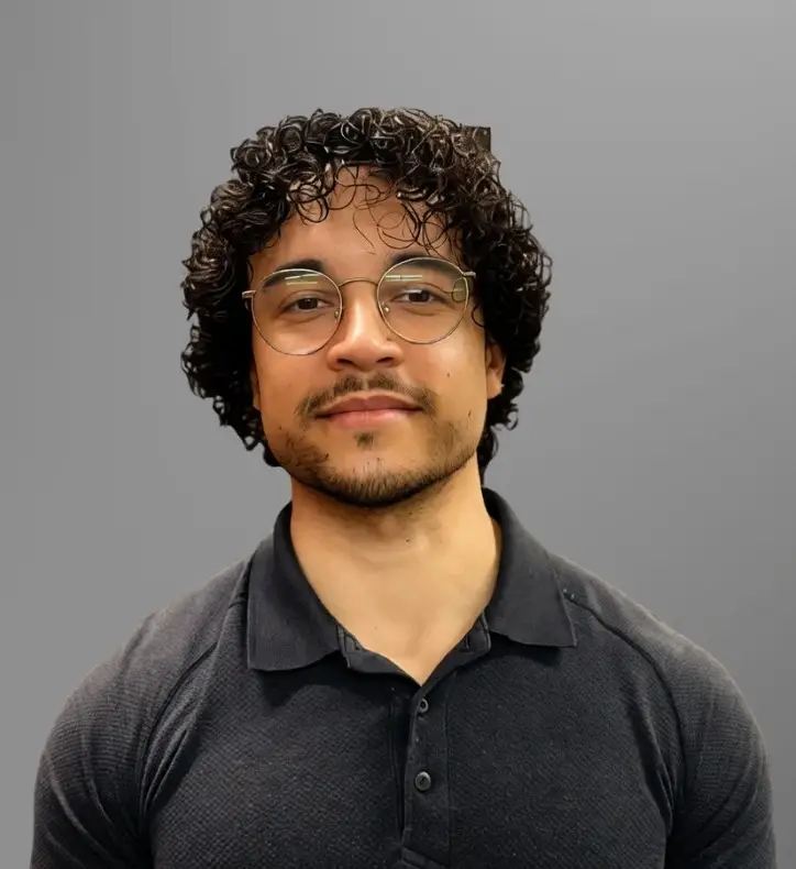 Luke Warner, a man with curly hair, glasses, and facial hair, is wearing a dark polo shirt and standing in front of a plain grey background.