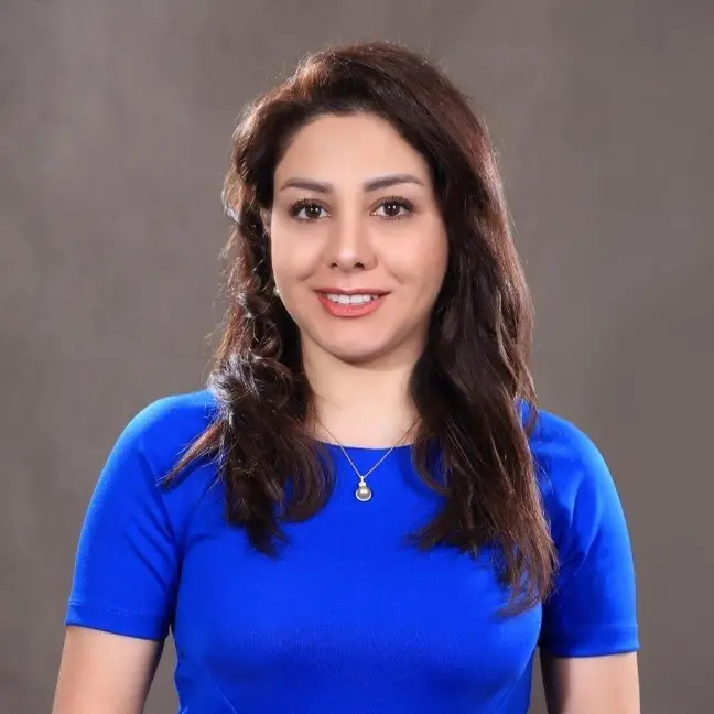 Woman with long dark hair wearing a bright blue short-sleeve shirt and a necklace, smiling at the camera in front of a plain gray background—a warm portrait by Luke Warner.